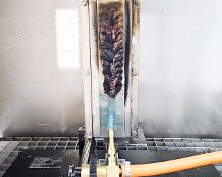 Forschungsprojekt »FireSafe-CLT«: Biobasierte Flammschutzmittel aus erneuerbaren Rohstoffen für den Brandschutz von Brettsperrholz Das Foto zeigt eine Laborapparatur: Auf einer vertikal eingespannten Metallplatte ist eine Klebstofffolie aufgebracht, die mithilfe einer Gasbrenner-Flamme von schräg unten beflammt wird.