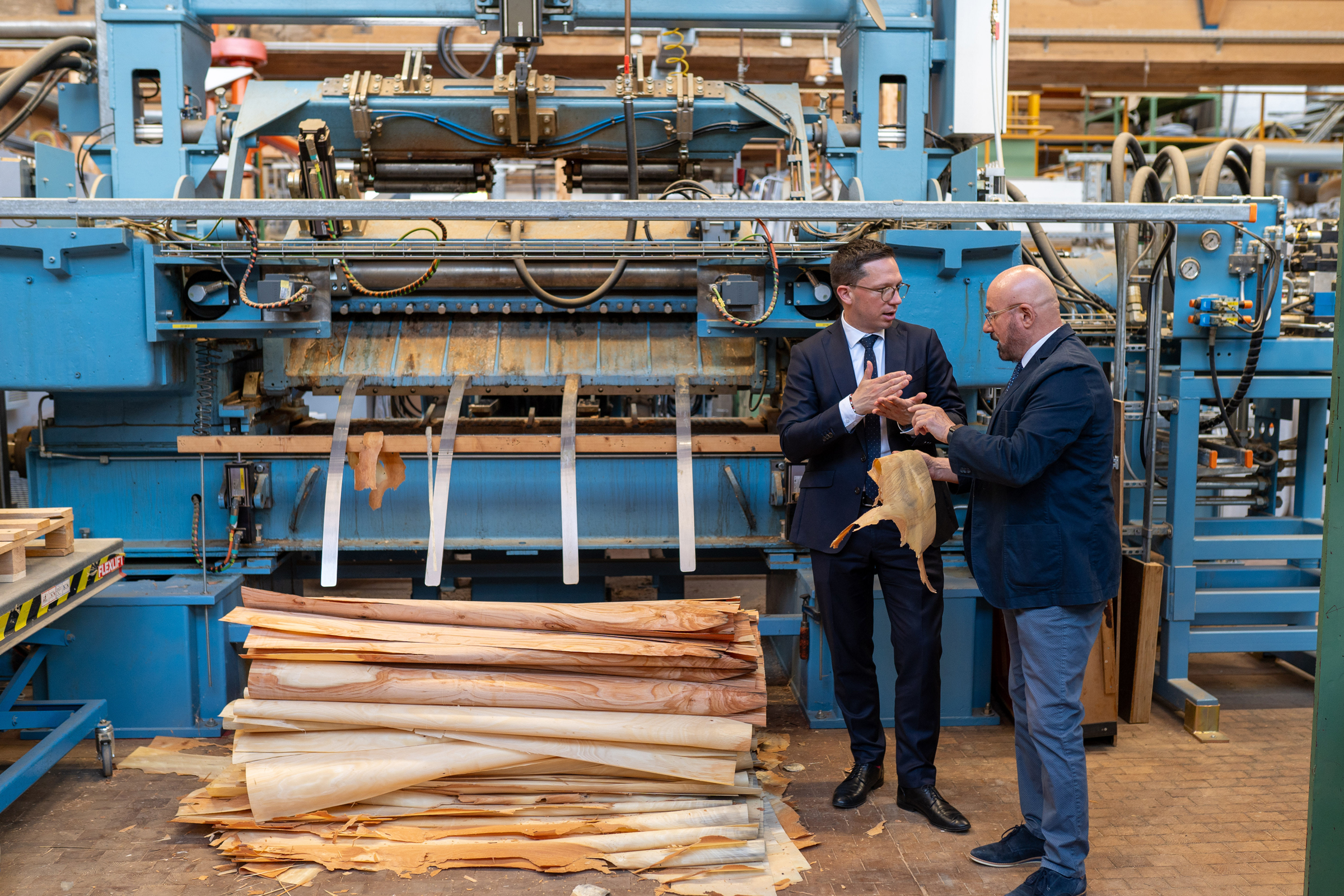Das Foto zeigt Minister Falko Mohrs und Institutsleiter Professor Kasal vor einer großen technischen Anlage, auf der gerade Holzfurnier hergestellt wird.