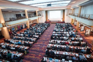 The photo shows a large room with rows of chairs on which people are sitting and a podium on which speakers are sitting.