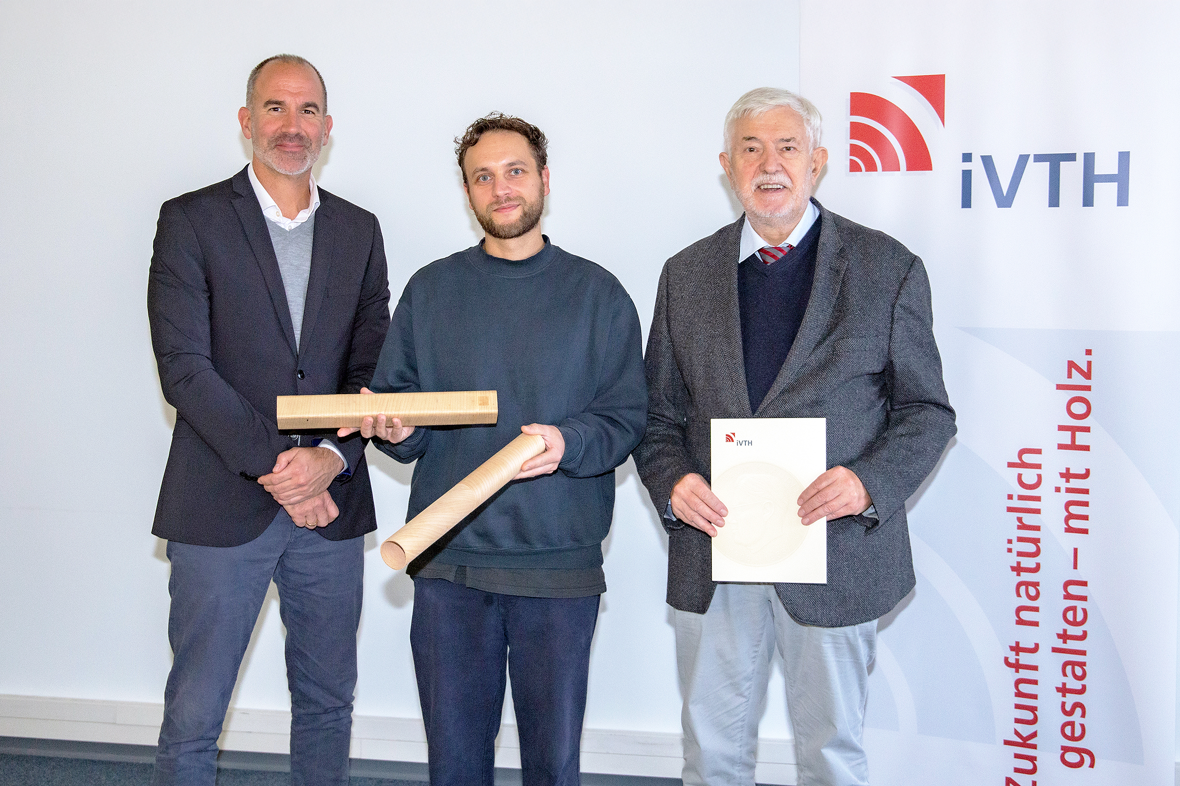 The photo shows three people. Prize winner Andreas Göbert holds hollow wooden profiles in his hand. Professor Dr. Marutzky holds a certificate in his hand.