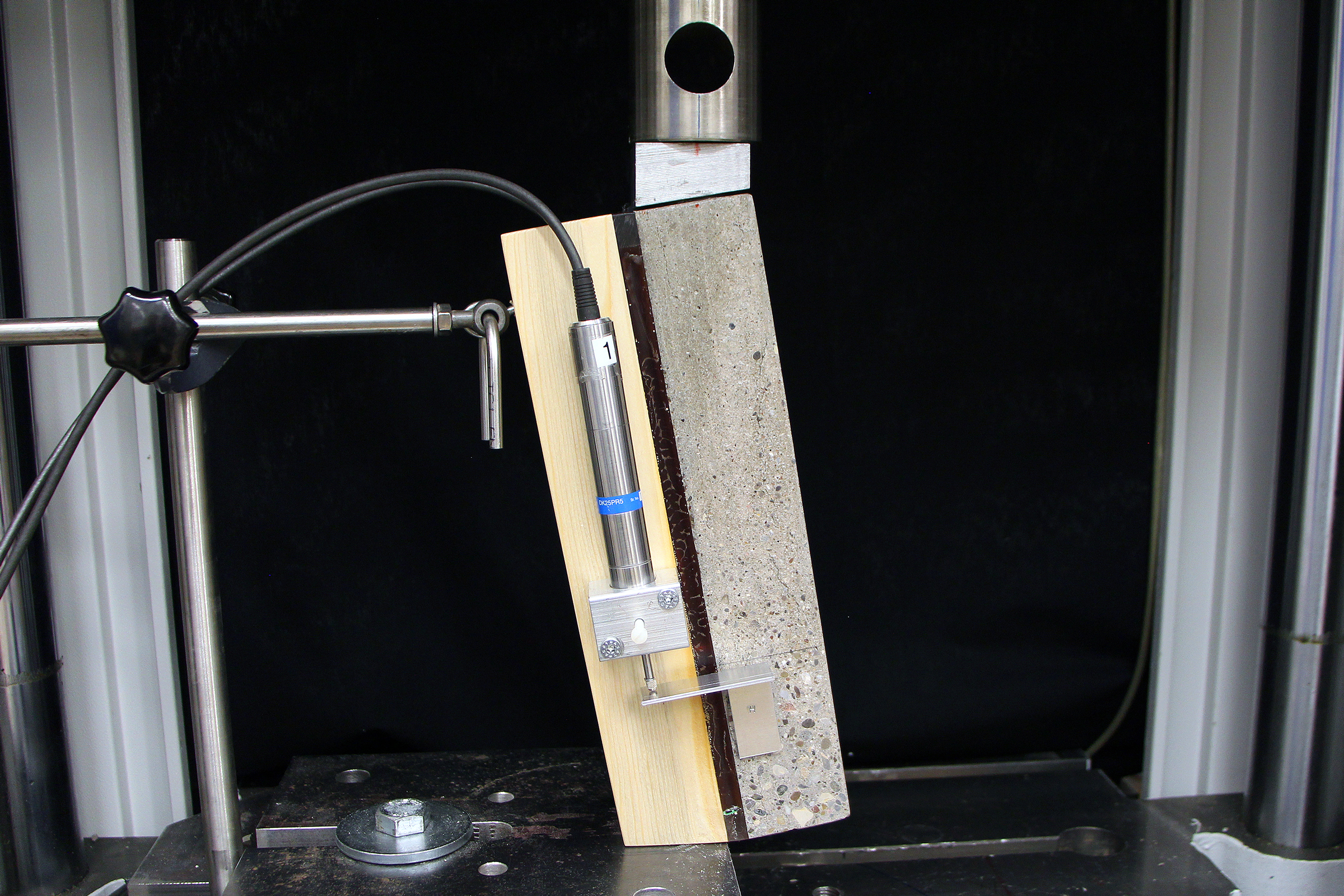 The photo shows a test specimen made of wood and concrete with a layer of adhesive in the testing device.