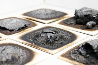 The photo shows several quadratic samples of a thin plywood board. On the upper surface of each of the samples of wood-based material is a mound of black-brown, foam-like material with a porous structure. The size and shape of the mound is different for each sample.