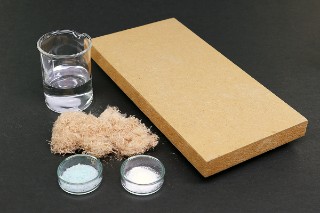 The photo shows a small piece of MDF, a small pile of wood fibers, a laboratory beaker containing a colorless liquid, a small laboratory dish containing pale-blue powder and a laboratory dish containing white powder.